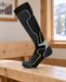 Black knee-high ski sock with grey panels and yellow outlines displayed on a wooden lodge bench.