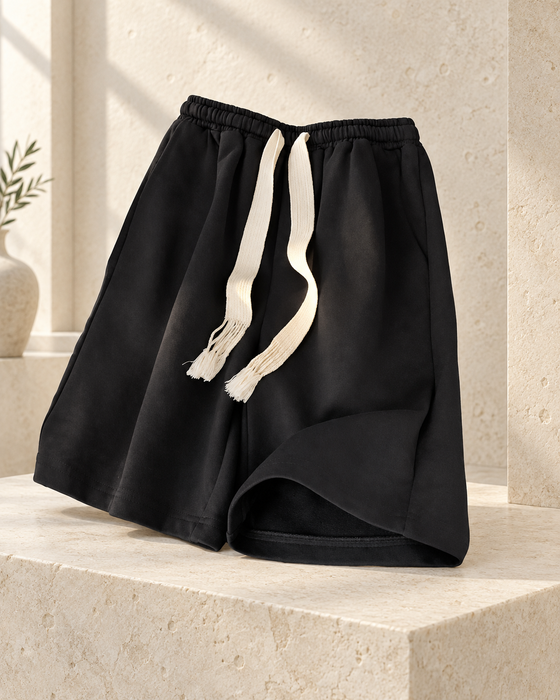 Black drawstring shorts with cream fringed cords displayed on pale limestone.