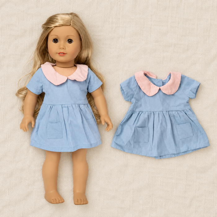 Blue doll dress with pink collar displayed on doll and separately on ivory linen