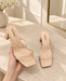 Beige square-toe sandals with thin crossing straps and block heels, shown as a pair in a dressing room.