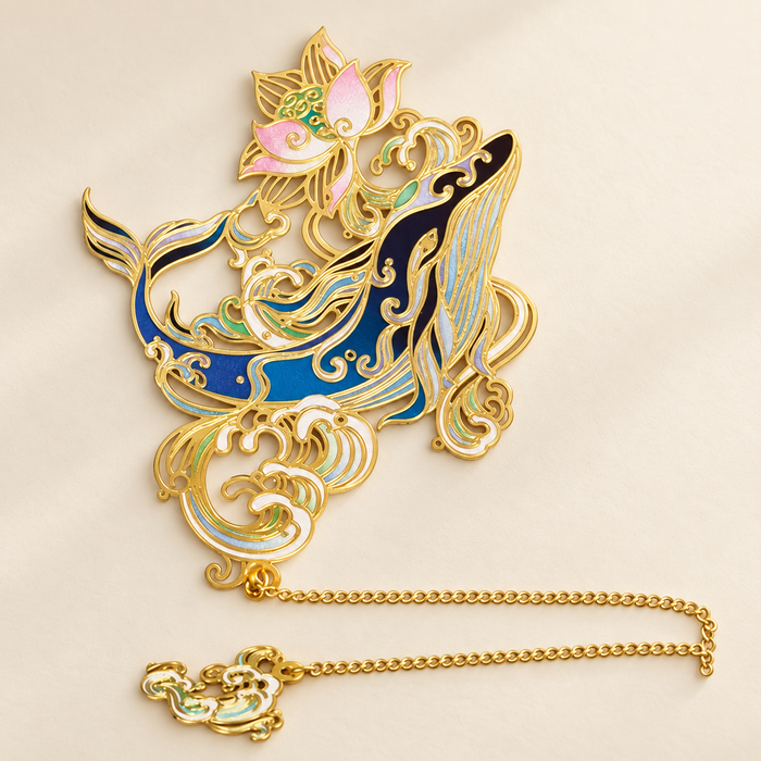 Blue and gold whale bookmark with pink lotus flowers and gold chain on ivory surface