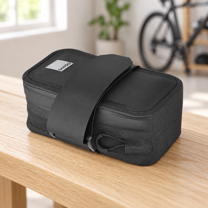 Black bicycle saddle bag with mounting strap on a wooden bench, with a bicycle blurred in the background.