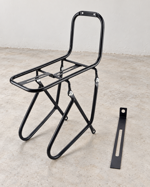 Black bicycle front carrier rack and a separate slotted mounting bracket on a pale concrete floor.
