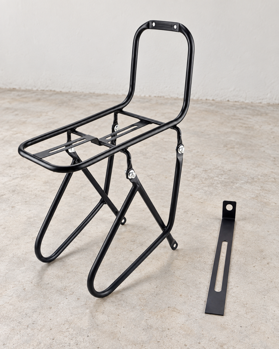 Black bicycle front carrier rack and a separate slotted mounting bracket on a pale concrete floor.