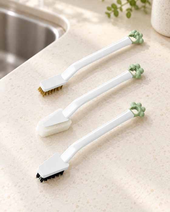 Three white cleaning brushes with green hanging loops on an ivory kitchen counter.
