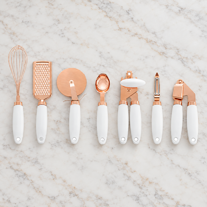 Seven rose-gold kitchen utensils with white handles arranged on a pale stone worktop.