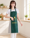 Woman wearing a green apron with grey side panels and yellow COOKING print in a bright ivory kitchen.