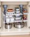 White two-tier kitchen rack arranged around pipes inside a cabinet.