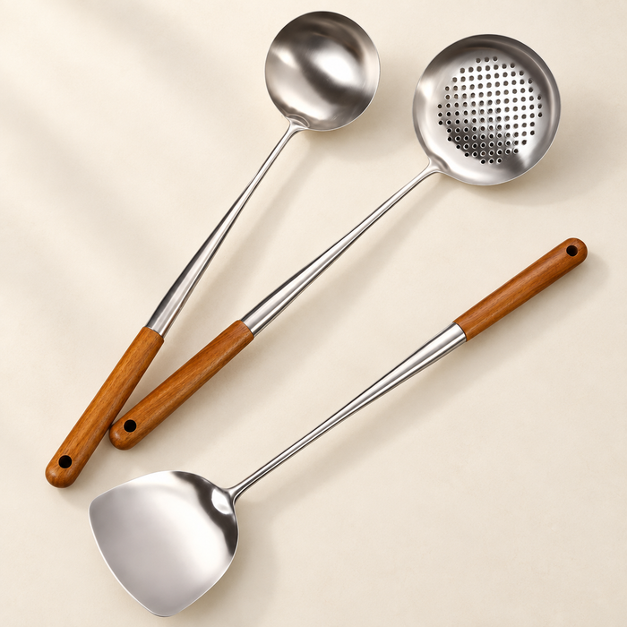 Three steel cooking utensils with wooden handles on an ivory surface.