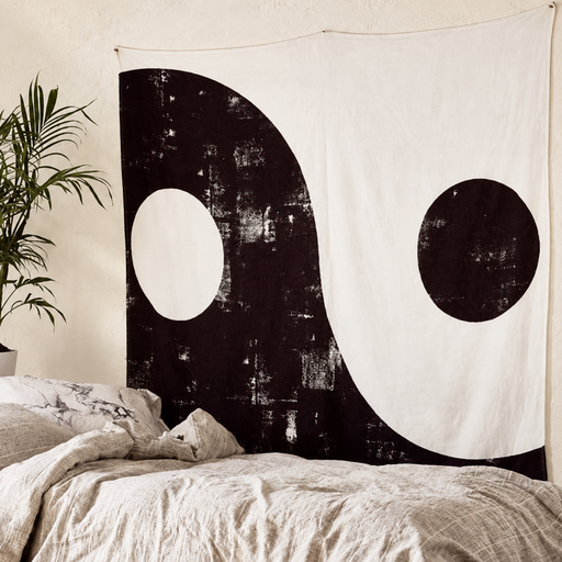Distressed black and white Yin-Yang wall tapestry above a bed beside a plant.