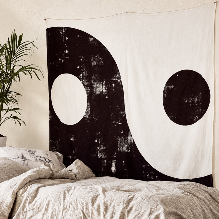 Distressed black and white Yin-Yang wall tapestry above a bed beside a plant.
