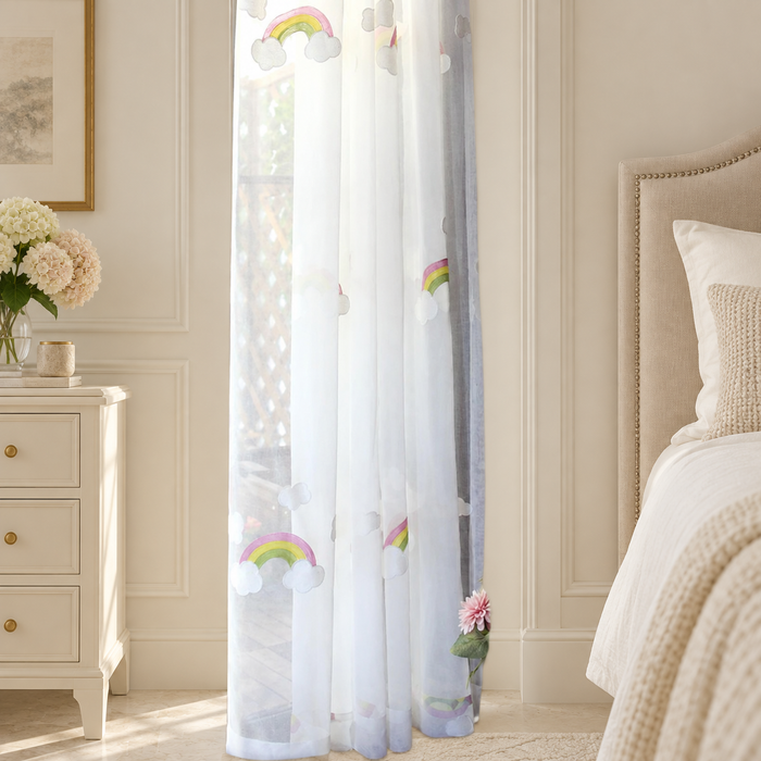 Sheer white curtain with pastel rainbow and cloud embroidery beside a bedroom window.