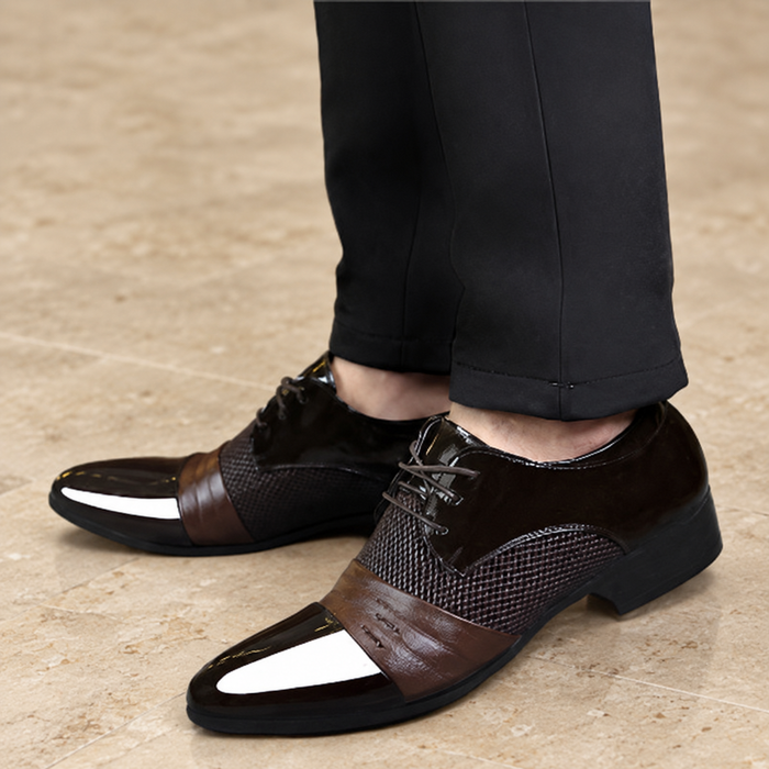 Brown and black glossy lace-up dress shoes worn with black trousers on a beige stone floor.