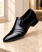 Black pointed-toe slip-on dress shoe with three gold bars and a V ornament on a pale stone surface.