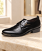 Black dress shoe with a honeycomb-textured panel and gold ornament on a pale stone bench.