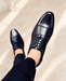 Navy lace-up dress shoes with cap-toe seams worn with dark trousers above a pale stone floor.