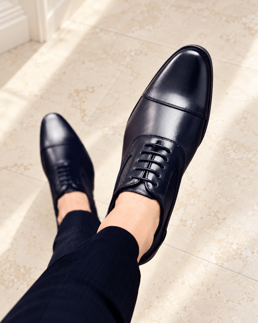 Navy lace-up dress shoes with cap-toe seams worn with dark trousers above a pale stone floor.