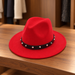 Red fedora with a black band and alternating silver studs on a wooden shelf.