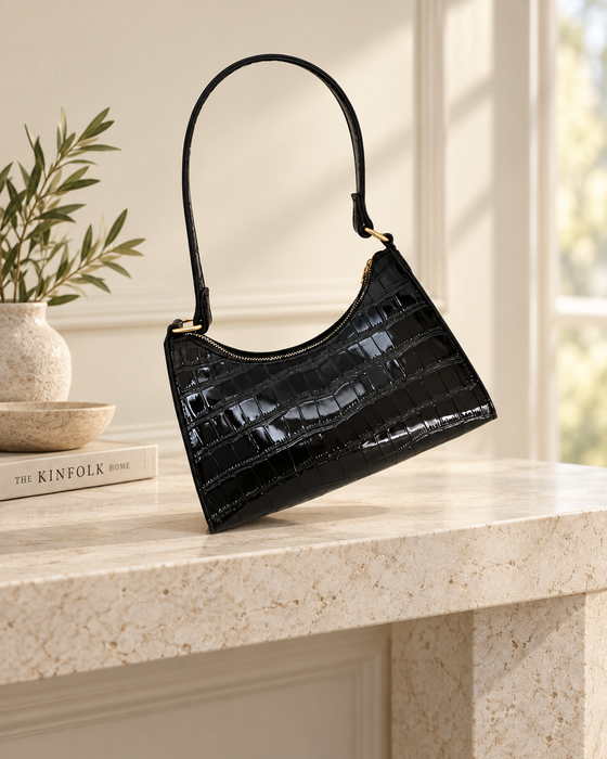 Black glossy crocodile-embossed shoulder bag with gold-tone strap links on a stone console.