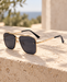 Gold metal-frame sunglasses with dark rectangular lenses resting on a sunlit travertine ledge.