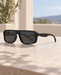 Matte black rectangular sunglasses with a silver hinge detail on a sunlit stone tabletop.