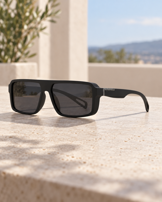 Matte black rectangular sunglasses with a silver hinge detail on a sunlit stone tabletop.