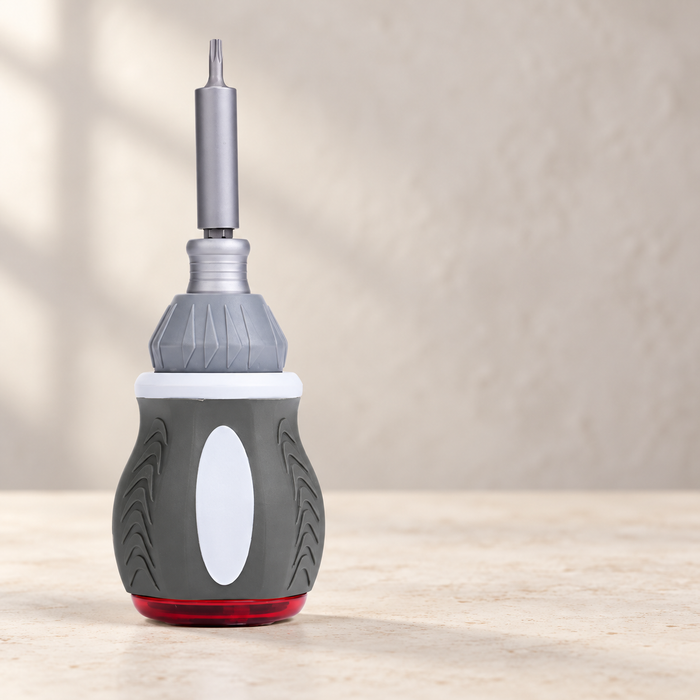 Grey ratchet screwdriver with white grip insert and red base on a pale worktable
