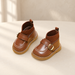 Brown children shoes with knit ankle cuffs, gold buckles and white stitching on an ivory surface.