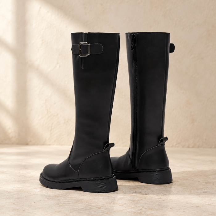 Tall black boots with a silver buckle and side zip on ivory stone